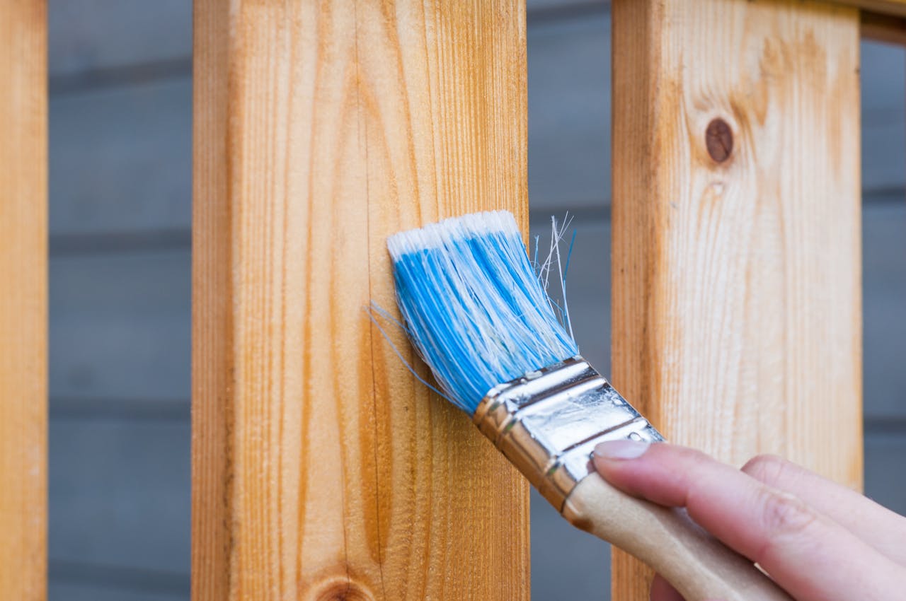 Servicios de mantenimiento EN SANT PERE PESCADOR: RENOVA HAUS A hand painting a wooden fence plank with a blue paintbrush outdoors. Perfect for DIY and renovation themes.